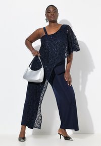 Mono de encaje marino con un solo hombro, que presenta intrincados patrones florales y una abertura lateral, combinado con un bolso plateado y tacones negros.