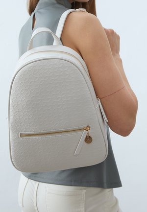 Femme portant un haut gris sans manches et un pantalon blanc, avec un sac à dos blanc à motif et une fermeture éclair dorée sur l'épaule.