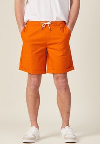 Pantaloni corti arancioni in cotone con vita regolabile tramite coulisse, tasche laterali e orli arrotolati, abbinati a scarpe da ginnastica bianche e una maglietta bianca semplice.