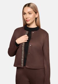 Brązowa kurtka z okrągłym kołnierzem, ozdobiona koralikowym wykończeniem i czarnymi guzikami. Teksturowany materiał dodaje subtelnej głębi.