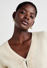 Beige cardigan med teksturfinish, med blondekant langs V-halsen og perleknaplukning. Korte ærmer, blødt materiale.
