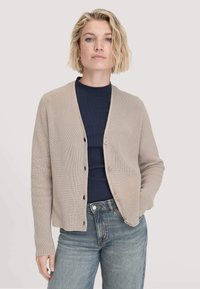 Frau mit kurzen blonden Haaren, die eine beige Knopfleiste-Kardigan über einem navyfarbenen Oberteil und blauen Jeans trägt und mit einer Hand in der Tasche posiert.