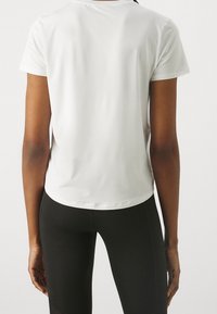Bílé sportovní tričko s krátkým rukávem, hladká textura, zakřivený lem, nošené s černými legínami, pohled zezadu. Žádné viditelné logo.