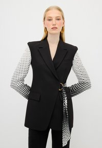 Blazer noir ajusté avec des manches à motifs contrastants en blanc et noir ; doté d'une ceinture avec une boucle circulaire et de poches minimalistes.