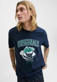 Ung mand med langt blondt hår og tatoveringer iført navy blue T-shirt med "ORIGINALS" og croissant grafik, ser til siden.