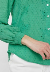 Blusa verde con texture, caratterizzata da ricami floreali, chiusura a bottoni e polsini elasticizzati. Accentuata da piccole punteggiature in rilievo.