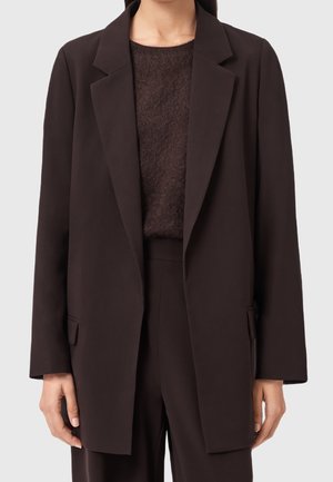 Femme portant un blazer marron foncé sur un pull marron foncé pelucheux et un pantalon assorti sur un fond clair uni.