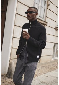 Melnā ribota zip-up jaka ar augstu apkakli, uzvilkta virs plāna krekla, saskaņota ar pelēkiem biksēm. Modelis tur kafijas krūzi ārā.