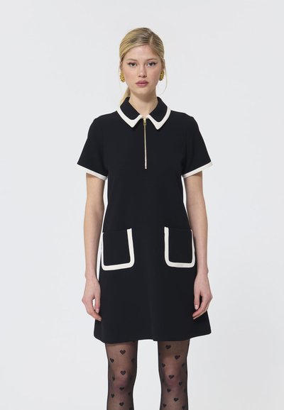 Abito nero con maniche corte, colletto bianco e finiture delle tasche, zip frontale, tessuto liscio, indossato con collant a motivo cuore.