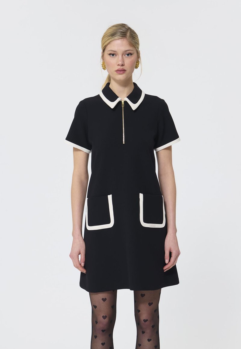 Vestido negro con mangas cortas, cuello blanco y acabados de bolsillo, cremallera frontal, textura suave, combinado con medias con estampado de corazones.