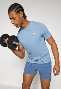 Lys blå kortermet treningsskjorte med Puma-logo, kombinert med blå shorts, som holder en svart dumbbell med metallgrep.