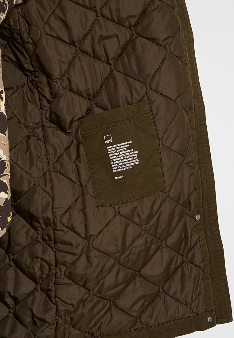 Gev quiltde bruine jas binnenkant, met een gestructureerd patroon en een zak met witte labeldetails. Het buitenmateriaal is corduroy.