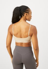 Sujetador deportivo beige con tirantes ajustables y banda de marca; tejido suave, diseño minimalista. Llevado con leggings oscuros de talle alto.