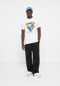White T-shirt with a colorful graphic print featuring a cartoon character, paired with black cargo pants and white sneakers. Light blue cap.