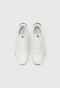 Witte Calvin Klein sneakers met veters, zwart logo op de tong en zwarte accenten op de zool, vanboven bekeken op een witte achtergrond.