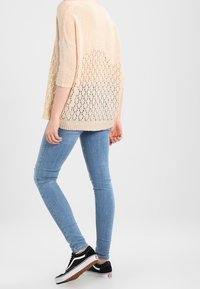 Krämfärgad stickad tröja med lös passform och urvändig mönster, kombinerad med ljusblå skinny jeans och svarta sneakers med vita accenter.