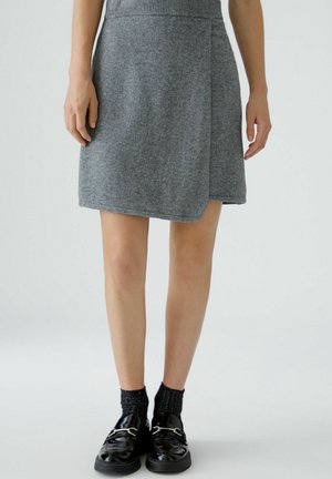Femme portant une jupe portefeuille en laine grise, des chaussettes noires pailletées et des mocassins noirs brillants, debout sur un fond clair uni.