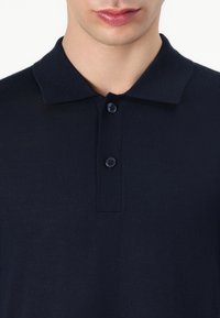 Polo shirt bleu marine foncé en tissu tricoté lisse. Doté d'un col classique et de trois boutons bleus au niveau de la patte de boutonnage. Design simple et minimal.