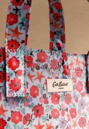 Ziedu auduma iepirkumu soma ar gaiši zilu fonu, uz kura attēloti sarkani un rozā ziedi un zvaigznes. Uz birkas ir uzraksts "Cath Kidston London." Izturīgas siksnas.