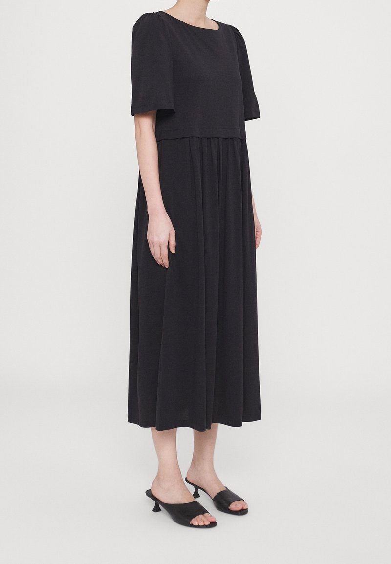 Femme portant une robe midi noire à manches courtes et des mules noires à bout ouvert, debout devant un fond clair uni.