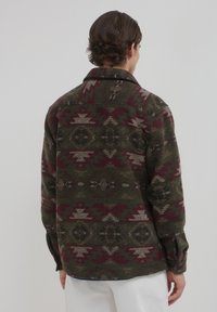 Uomo che indossa una camicia a maniche lunghe con bottoni, caratterizzata da motivi geometrici verdi, bordeaux e beige, visto di spalle su uno sfondo semplice.