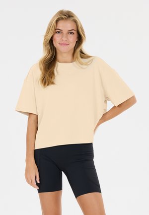 Ung kvinde med blondt hår iført en løs beige kortærmet top og tætsiddende sorte cykelshorts står med den ene hånd på hoften.
