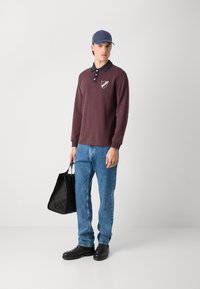 Polo de culoare maroon cu mâneci lungi, guler și emblemă de culoare navy, asortat cu blugi de un albastru deschis și pantofi negri. O geantă tote neagră este ținută în una dintre mâini.