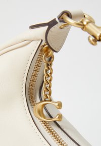 Borsa a mano in pelle crema con zip dorata e un accento a catena, con un charm dorato unico attaccato. Texture liscia con cuciture minime.