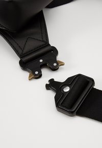 Mustan nahkavyön mustalla metalliosalla ja kultaisella yksityiskohdalla. Varustettu kiinnitysmechanismilla ja teksturoidulla nylon-osalla.