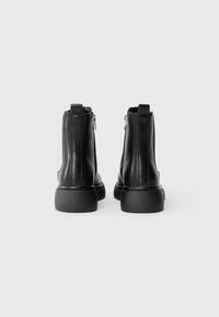 Botas de tobillo de cuero negro con una textura suave, suela de goma plana y cremalleras metálicas en la parte trasera, con elementos de diseño minimalistas.