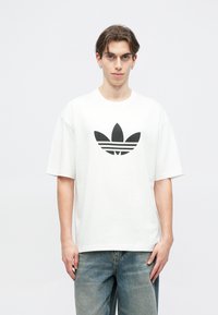 Biele tričko s krátkymi rukávmi vyrobené z bavlny, s veľkým čiernym logom trefoilu Adidas na prednej strane, v kombinácii s modrými džínsami.