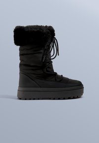 Czarny ocieplany but z kołnierzem z sztucznego futra, pikowaną górą, płaskimi sznurówkami i grubą gumową podeszwą z fakturowanym bieżnikiem.