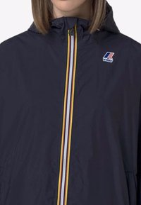 Veste coupe-vent marine avec un col montant, dotée d'une fermeture éclair centrale avec des rayures rouges, jaunes et blanches, et d'un petit logo sur la poitrine.