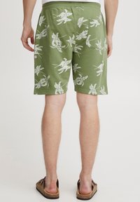 Green shorts made of fabric with white tropical leaf patterns, featuring a back pocket and an elastic waistband for comfort.