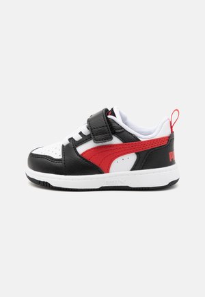 Baskets basses enfant en blanc, noir et rouge avec une bride Velcro, une bande rouge sur le côté et une languette sur une semelle blanche sur fond clair.
