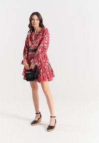 Vestito rosso a fiori con maniche a volant, gonna fluida e scollatura a V; abbinato a una borsa nera e sandali con zeppa e cinturino alla caviglia.