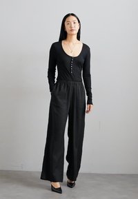 Black long-sleeve bodysuit with a scoop neckline and button detail, paired with high-waisted, wide-leg trousers. Both garments are solid black.