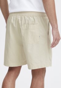 Shorts din bumbac bej cu un tiv elastic, având un singur buzunar pe spate și un model texturat subtil. Marginile sunt drepti.