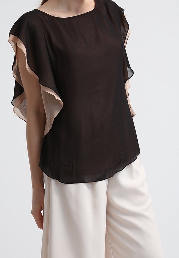 Haut noir transparent avec des accents beige fluides, encolure arrondie et manches courtes. Le tissu tombe doucement sur un pantalon de couleur claire.