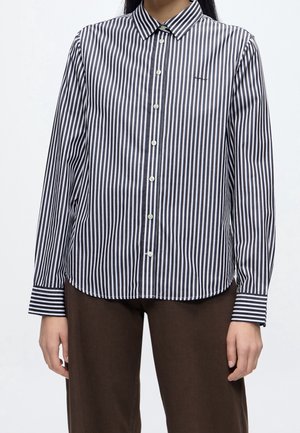 Personne portant une chemise à rayures verticales noires et blanches avec boutons, manches longues, et un pantalon marron sur un fond uni.