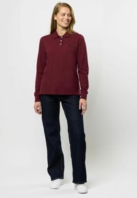 Bordeauxrood polo shirt met lange mouwen, een kraag en drie knopen, gecombineerd met donkerblauwe jeans en witte sneakers. Eenvoudig logo detail op de shirt.