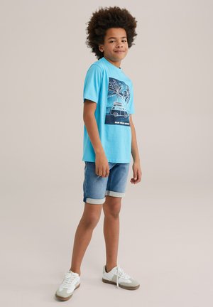 T-shirt en coton bleu clair avec un imprimé graphique de palmiers et d'une camionnette, associée à un short en denim avec des ourlets roulés et des baskets blanches.