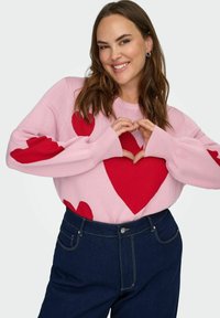 Růžový pletený svetr s červenými vzory srdce, volnými rukávy a kulatým výstřihem, kombinovaný s tmavými džínami.