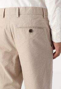 Beige textured trousers with a subtle pattern, featuring a back pocket with a brown button and a waistband with belt loops.