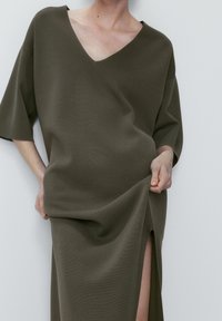 Olive green, knee-length dress with a V-neck, wide sleeves, and a side slit. Made from textured fabric, featuring a loose fit.
