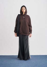 Sweatshirt castanha com capuz e bolso frontal; combinado com calças largas em cetim preto com riscas laterais brancas. O modelo segura uma mala preta.