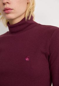 Primo piano di una persona che indossa un dolcevita a maniche lunghe viola a coste con un piccolo logo ricamato sul petto.