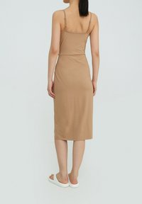 Robe midi beige à fines bretelles et silhouette ajustée, confectionnée en tissu lisse et extensible, présentée vue de dos. Remarquable par sa simplicité.