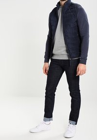Chaleco acolchado azul marino sobre una sudadera gris, combinado con jeans denim oscuros, doblados en los tobillos, y zapatillas blancas. Diseño minimalista, sin estampados.
