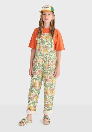 Pige iført orange t-shirt, blomstrede grønne og gule overalls, matchende sandaler og en hvid og grøn kasket med tekst på fronten.